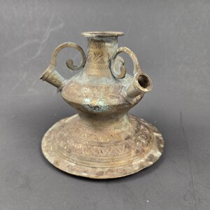 May include: A weathered brass vessel featuring a wide, flat base, a central neck, and two symmetrical side spouts with curved handles. The metal surface displays a tarnished patina with intricate, engraved geometric patterns throughout the body.