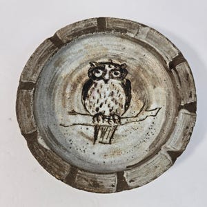 May include: A round ceramic tray with a textured, mottled gray and brown finish. The center features a hand-drawn illustration of an owl perched on a branch in dark brown ink. The wide, raised rim has a segmented, rustic appearance with dark brown accents.