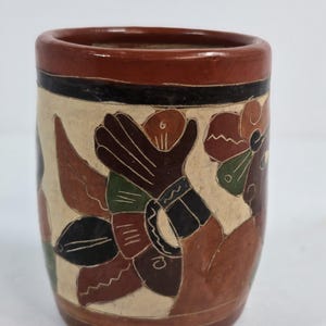 Puede incluir: Una taza cilíndrica de cerámica pintada a mano con un diseño floral. La taza presenta un borde y una base de color marrón rojizo, con una banda central de color crema. El diseño floral incluye elementos marrones, verdes y negros.
