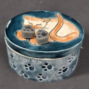 Puede incluir: Una caja redonda de cerámica con tapa y esmalte azul, con un gato naranja pintado en la tapa y dos pequeños pomos grises en forma de huella. El lateral está decorado con huellas de color azul oscuro y el texto MEOW 1979 en relieve.