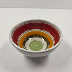 Puede incluir: Un pequeño cuenco de cerámica blanca con un diseño concéntrico vibrante. El interior presenta anillos rojos, naranjas, negros y verdes. El borde del cuenco tiene una banda roja, y la estética general es colorida y decorativa.