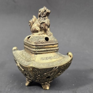 May include: A metallic gold-toned incense burner featuring a square lid topped with a sculpted guardian lion figure. The body of the vessel is decorated with intricate embossed dragon motifs and geometric patterns, supported by three small ornate feet.