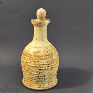 Puede incluir: Una jarra de cerámica con cuello estrecho y tapón esférico. El recipiente presenta un esmalte de color crema con patrones de manchas marrones y líneas texturizadas horizontales que envuelven el cuerpo, terminando en una base ensanchada.