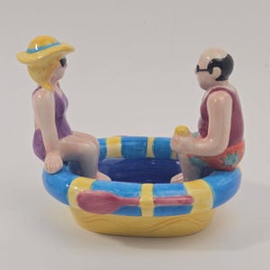 May include: A ceramic bowl shaped like a small boat with two figures sitting on the edges. One figure wears a yellow hat and purple swimsuit, while the other wears dark sunglasses and patterned swim trunks. The boat features blue and yellow stripes.