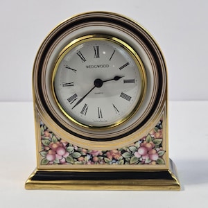 May include: A Wedgwood quartz mantel clock with an arched ceramic frame featuring a black and gold border. The white clock face displays black Roman numerals and the text Wedgwood and Quartz. The base is decorated with a colorful floral pattern.