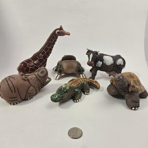Puede incluir: Una colección de seis figuras de animales de cerámica que incluye una jirafa marrón, un hipopótamo texturizado, una rana verde, una vaca blanca y negra, un cocodrilo verde y una tortuga marrón, dispuestos alrededor de una moneda para escala.