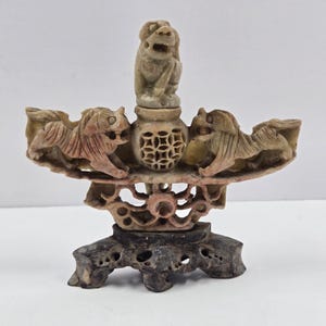 Puede incluir: Escultura de piedra tallada con dos figuras de leones estilizados que flanquean un jarrón central con una figura de león en la parte superior. La escultura tiene una base de varios niveles y está hecha de piedra marrón y beige.