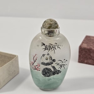 Puede incluir: Una botella de rapé de vidrio transparente con un panda pintado a mano en blanco y negro, sentado entre tallos de bambú y flores de cerezo rosas. La botella tiene forma redondeada, un cuello plateado y un tapón de piedra redondo y moteado.