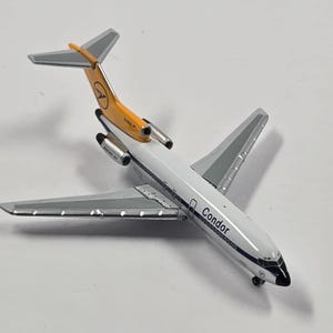 Może przedstawiać: Miniaturowy model samolotu, z białym kadłubem z napisem "Condor". Ogon jest żółty i szary, z logo. Skrzydła są srebrne. Model to Boeing 727.