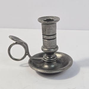 Puede incluir: Un pequeño candelabro de metal color estaño con base circular, un soporte cilíndrico central con dos ranuras horizontales y un asa lateral que incluye un apoyo para el pulgar y un anillo circular para sujetarlo con el dedo.