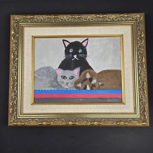 May include: A framed painting featuring three cats: a black cat in the center, a gray cat on the left, and a brown cat on the right. The cats are positioned above horizontal stripes of blue, red, and black. The painting is in a gold ornate frame.