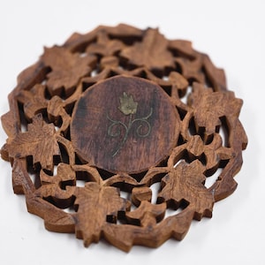 May include: A round, intricately carved wooden coaster with a dark brown finish. The coaster features a central circular design with a floral inlay and is surrounded by a detailed leaf pattern. The coaster is approximately 15 cm in diameter.