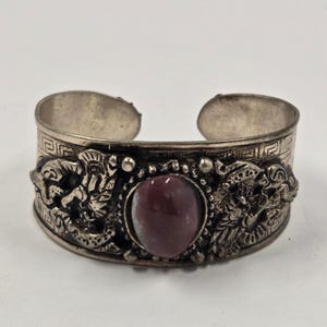 Puede incluir: Brazalete de puño plateado con una piedra central ovalada de color granate. El brazalete presenta motivos detallados de dragones a ambos lados de la piedra y un patrón geométrico en el borde superior. Una pieza llamativa.