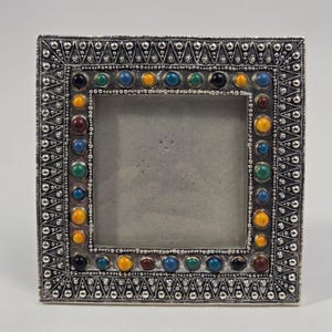 May include: A square silver-toned picture frame featuring an intricate border of embossed triangular patterns and small metal beads. The inner edge is decorated with a single row of colorful, smooth, round cabochon stones in yellow, blue, green, and dark red.