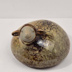 Puede incluir: Una figura de oveja de cerámica con un cuerpo redondeado en forma de cúpula que presenta un esmalte moteado en tonos marrón y bronceado. La cabeza es lisa y de color claro con acentos marrón oscuro alrededor de las orejas y los ojos.