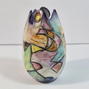 Puede incluir: Un jarrón de cerámica en forma de huevo con borde festoneado, que presenta un patrón abstracto pintado a mano de formas geométricas en tonos morado, azul, amarillo, verde y naranja, delineado con líneas negras gruesas sobre fondo blanco.