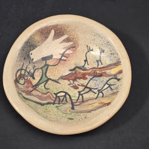 May include: A round ceramic bowl with a beige base featuring a hand-painted scene in black, green, red, and yellow. The design depicts a large white hand-like shape above a stick figure and several four-legged animal silhouettes on a landscape.