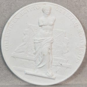 May include: A circular white plaster medallion featuring a central relief sculpture of a standing armless figure draped in fabric. The text COLLECTIS EX EGYPTO GRAECIAQ MONUMENTIS is embossed along the curved border, surrounding the central figure.