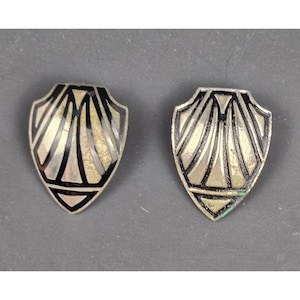 Puede incluir: Un par de pendientes de metal en forma de escudo con acabado plateado e incrustaciones de esmalte negro. El diseño presenta líneas radiantes y patrones geométricos que forman un motivo en forma de abanico en la superficie de cada pendiente.