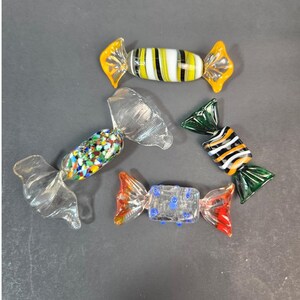 Puede incluir: Cuatro esculturas de vidrio con forma de caramelos envueltos. Una presenta rayas amarillas, blancas y negras. Otra tiene un patrón de confeti multicolor. Una tercera muestra rayas negras, blancas y naranjas con extremos verdes. Una es transparente con puntos azules.