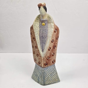 May include: A ceramic figurine of a stylized figure wearing a textured, rust-colored coat with a repeating dark circular pattern over a lavender-colored garment. The figure has a cream-colored collar, a yellow circular accent, and a blue-gray base.