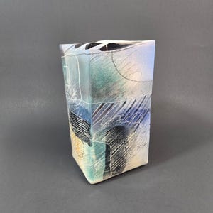 Puede incluir: Un jarrón de cerámica rectangular con una superficie multicolor con degradados de azul, verde azulado y crema. El exterior está decorado con intrincados trazos de líneas blancas y negras, incluyendo sombreados cruzados, garabatos y líneas finas.