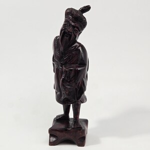 Peut inclure: Une figurine en bois sculpté brun foncé représentant un personnage debout avec une longue barbe, portant des robes traditionnelles et une coiffure en chignon. La figurine est posée sur un socle carré et présente des textures détaillées.