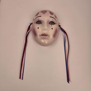 Puede incluir: Una máscara facial de cerámica brillante con acabado pálido, pintada con formas de corazón moradas en la frente, sombra de ojos morada oscura, detalles de lágrimas azules bajo los ojos y pequeños corazones rojos en las mejillas, con largas cintas burdeos y azules.