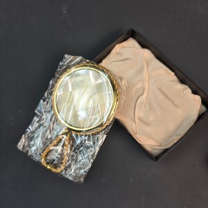 Puede incluir: Un espejo de mano redondo con marco de metal dorado y mango, que presenta una incrustación de mosaico de azulejos de nácar blanco iridiscente. El espejo está envuelto en plástico transparente y descansa junto a una bolsa de tela beige.