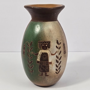 May include: A ceramic vase with a flared rim and a rounded body featuring a dark brown neck. The body displays a green and beige gradient finish with a dark brown etched figure of a person wearing a sun emblem, flanked by two vertical plant motifs.
