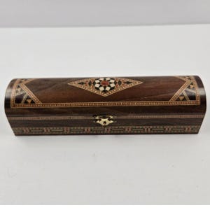 Puede incluir: Una caja de madera oblonga de color marrón oscuro con intrincados patrones geométricos incrustados. La caja presenta una tapa con bisagras y un cierre metálico decorativo. Los diseños incrustados incorporan acentos blancos, rojos y negros.