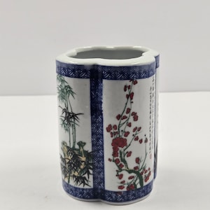 Puede incluir: Un jarrón de cerámica blanco con una forma única, con bordes geométricos azules. El jarrón está decorado con imágenes pintadas de bambú y ramas florecientes rojas. El jarrón mide aproximadamente 13 cm de altura.