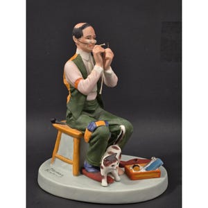 Puede incluir: Una figura de porcelana de una persona con gafas, camisa clara y chaleco verde, sentada en un taburete de madera mientras enhebra una aguja. Un gato pequeño está de pie cerca de los pies, junto a una caja naranja con herramientas de costura.