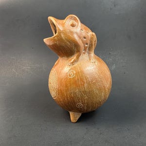 May include: A brown ceramic vessel shaped like a frog with its mouth open. The round body features etched circular and cross-hatched patterns. The sculpture stands on three small feet and has a smooth, matte finish with visible surface texture.