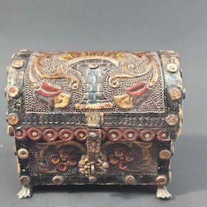 May include: A small, rectangular chest with a domed lid, featuring intricate embossed floral and geometric patterns in shades of black, brown, red, and gold. The box has a central metal latch and stands on four small, light-colored claw-shaped feet.