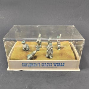 Puede incluir: Un conjunto de seis pequeñas figuras de circo de metal plateado dispuestas sobre una base de espuma color canela dentro de una caja de plástico transparente. La caja tiene el texto azul CHILDREN'S CIRCUS WORLD impreso en la base.
