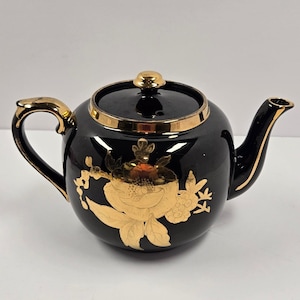 Puede incluir: Tetera de cerámica negra con diseño floral dorado y ribetes dorados. La tetera tiene un asa y un pico curvos, y una tapa con un pomo dorado. El diseño floral está en la parte delantera.