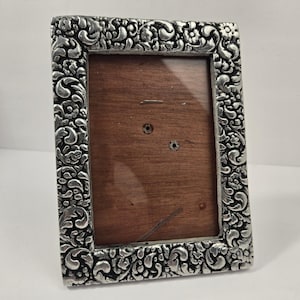 Puede incluir: Un marco de fotos rectangular con un borde de metal plateado que presenta un intrincado patrón en relieve de flores y volutas. El centro del marco contiene una pieza de madera de color marrón oscuro lisa protegida por un cristal.