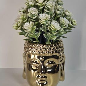 May include: A gold-coloured Buddha head vase filled with artificial white and green flowers. The vase has a glossy finish and detailed facial features. The flowers are clustered, creating a decorative arrangement.