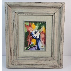 Puede incluir: Una pintura enmarcada que presenta un retrato estilizado con pinceladas abstractas y vibrantes en tonos verde, amarillo, rojo y azul. La obra está colocada dentro de un marco de madera desgastada de color claro con un paspartú de tela beige.