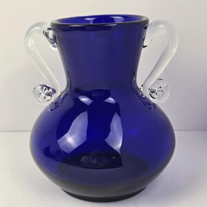 Puede incluir: Un jarrón de cristal azul cobalto con dos asas claras y curvas. El jarrón tiene un cuerpo redondeado y una abertura acampanada. Las asas están unidas cerca de la parte superior y se curvan hacia afuera, terminando en un remolino decorativo.