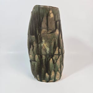 Puede incluir: Un jarrón de cerámica alto y cilíndrico con una forma orgánica irregular. La superficie presenta un esmalte moteado en tonos marrón oscuro, verde salvia y beige, con patrones de goteo verticales que recorren el cuerpo del recipiente.
