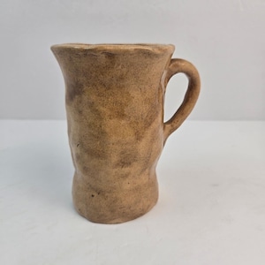 Puede incluir: Una taza de cerámica de color tostado con una superficie texturizada e irregular y un asa. La taza presenta un acabado moteado en tonos marrón y beige con un borde acampanado y un cuerpo cilíndrico que se estrecha ligeramente hacia la base.