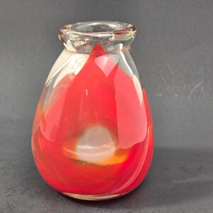 Puede incluir: Un pequeño jarrón de vidrio en forma de lágrima con un borde y cuerpo transparentes, que presenta patrones de color rojo y blanco vibrantes y arremolinados en su interior. El vidrio tiene un acabado liso y pulido con una base ancha.