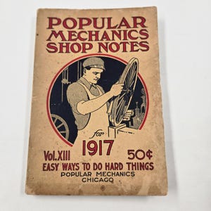Puede incluir: Portada de libro vintage "Popular Mechanics Shop Notes". La imagen muestra a un hombre trabajando en una rueda de bicicleta dentro de un círculo rojo. El texto incluye "Vol.XIII 1917" y "Easy Ways to Do Hard Things". El libro es de color beige con texto rojo y negro.