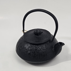 Op de afbeelding: Een zwarte gietijzeren theepot met een getextureerd oppervlak en reliëfpatronen van bladeren. De theepot heeft een gebogen handvat, een korte tuit en een rond deksel met een kleine, afgeronde knop. Een dun goudkleurig touwtje is aan het handvat bevestigd.