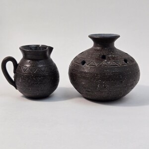 May include: A dark brown ceramic creamer with a handle and a matching round ceramic vase featuring a series of small circular openings around the top. Both pieces have a textured, speckled finish and a decorative band of incised triangle patterns.