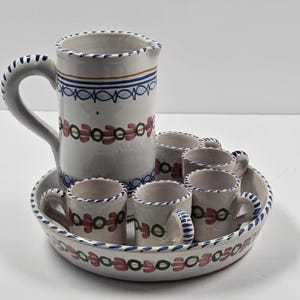 Puede incluir: Juego de jarra y tazas de cerámica blanca. La jarra tiene un asa grande y está decorada con motivos florales azules, marrones y rojos. Las tazas están dispuestas en una bandeja a juego, también decorada con los mismos motivos.