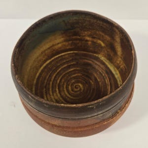 Puede incluir: Un cuenco de cerámica redondo con un borde marrón oscuro y una base de color marrón rojizo más claro. El interior presenta un patrón en espiral visible grabado en la arcilla, con tonos marrones y bronceados moteados en la superficie.