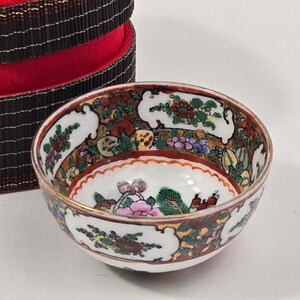 Puede incluir: Un pequeño cuenco de porcelana redondo con base blanca y complejos patrones florales multicolores en rojo, verde, rosa y dorado. El cuenco tiene un borde festoneado y se sitúa frente a un recipiente cilíndrico de color marrón oscuro y rojo.